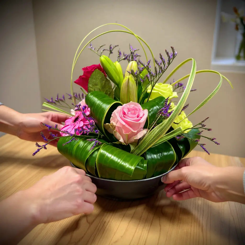 Bloemstuk halve bol color: een ronde bloemarrangement met roze roos, gele en roze bloemen, groene bladornamenten en paarse lavendel, vastgehouden door meerdere handen.